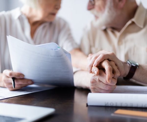 Cropped view of senior couple holding hands near documents, laptop and credit card on table